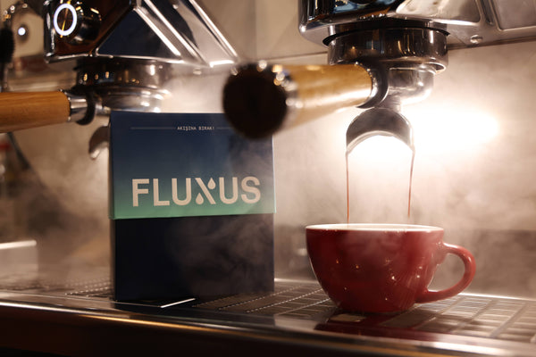 Espresso makinesinden kırmızı fincana akan espresso ile "FLUXUS" yazılı kahve paketi görülen bir kafe ortamı.