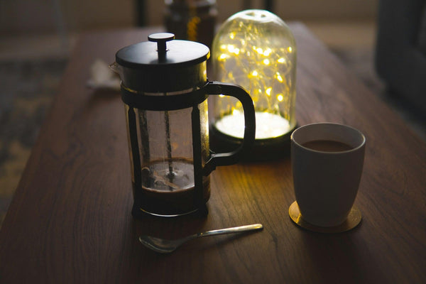 ahşap masa üstünde ışıklı lamba ve frech press ile demlenmiş filtre kahve